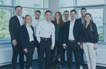 Team-IT-Projekt-und-Prozessmanagement Eight coworkers in business attire pose in a bright office with large windows; a black dog sits at their feet.