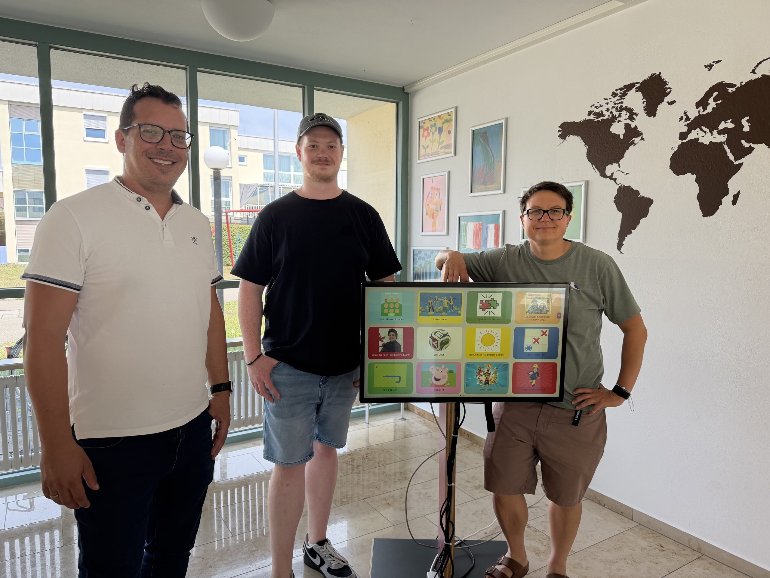 DORINO_gemeinsam_entwickelt-Dominikus-Ringeisen-Werk-SONNTAG-IT-Solutions Three adults pose with a large interactive touchscreen display in a bright room, with a world map mural on the wall behind them.