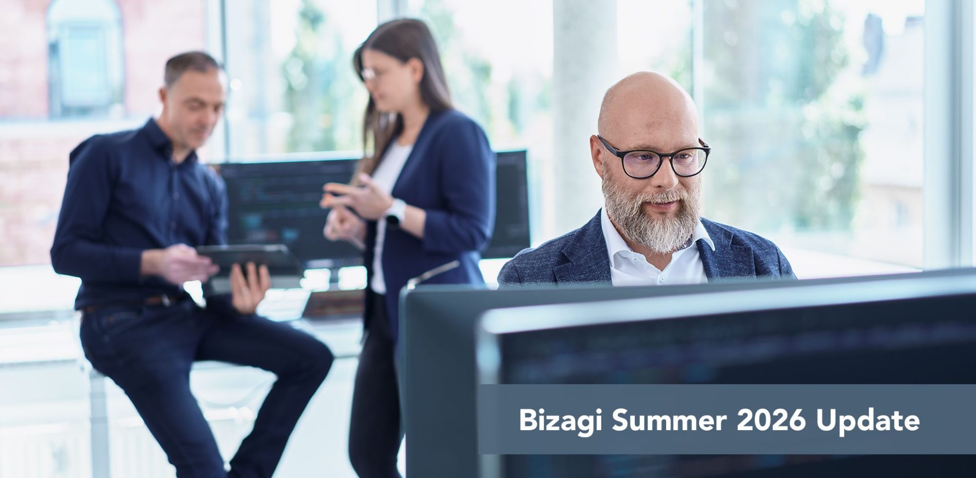 SITS-bizagi-summer-update Foreground: man with glasses at a computer in a bright office, two coworkers collaborate in the background, banner reads 'Bizagi Summer 2026 Update'.