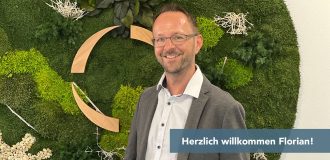 Smiling man in a blazer and glasses stands in front of a green moss wall with decorative wooden arcs behind him, and a 'Herzlich Willkommen Florian' banner partially visible.