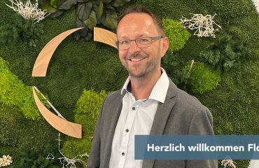 Smiling man in a blazer and glasses stands in front of a green moss wall with decorative wooden arcs behind him, and a 'Herzlich Willkommen Florian' banner partially visible.